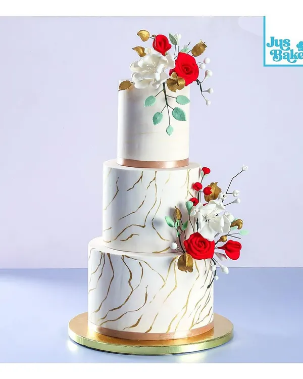 This three-tier wedding cake features a striking marble effect with gold veining, accented by handmade red and white sugar flowers. It's a modern and artistic choice for a memorable wedding day.