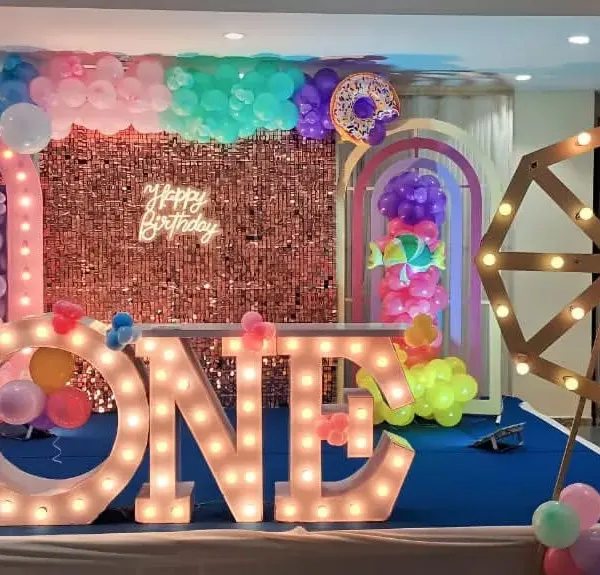 A grand 'Sweet One' themed first birthday party setup. This features a large rose gold shimmer wall, custom light-up 'ONE' marquee letters, a decorative ferris wheel, and multiple balloon arches for a truly memorable celebration.