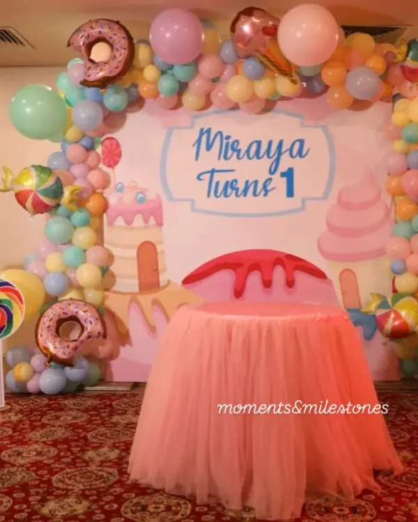 A sweet-themed cake-cutting area for Miraya's first birthday. The backdrop is surrounded by a pastel balloon arch with donut and candy-shaped foil balloons, and the cake table is dressed in a pink tutu skirt.