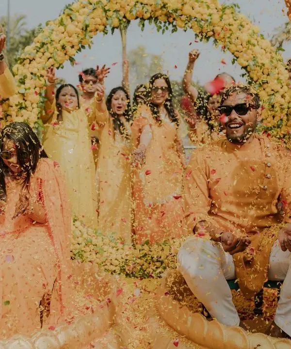 A shower of flower petals marks the climax of a fun-filled Haldi. This wide shot captures the collective joy of family and friends celebrating the couple under a beautiful floral arch.