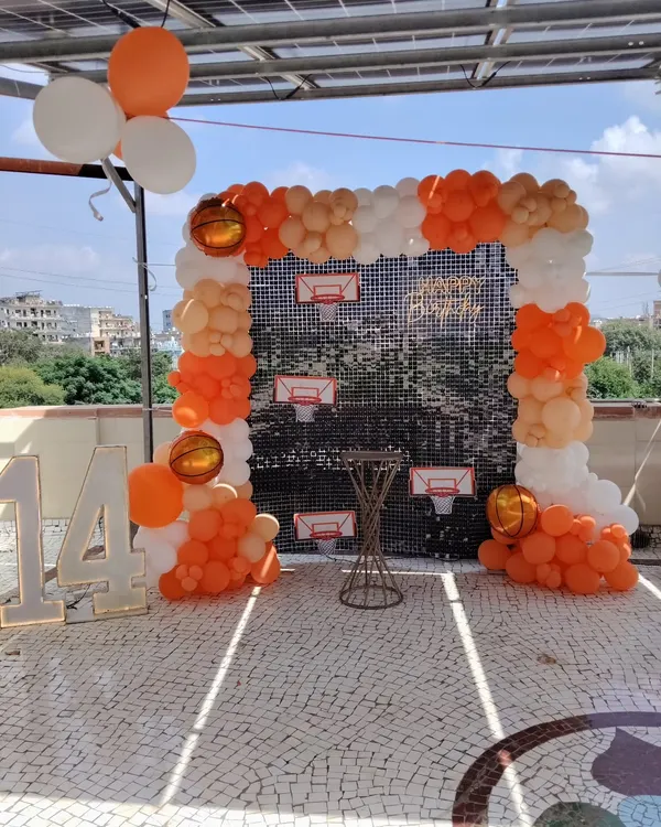 A basketball-themed 14th birthday on a sunny terrace. The setup includes an orange and white balloon arch against a sequin backdrop and large marquee numbers.