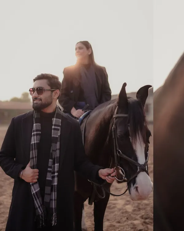 A stylish equestrian-themed shoot. The groom leads the horse while the bride sits elegantly, both dressed in chic black winter wear, creating a sophisticated and unique pre-wedding portrait.
