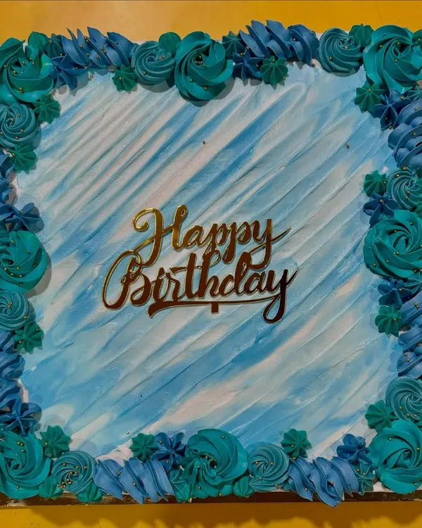 A beautiful square birthday cake with a watercolor-effect blue frosting. The border is decorated with hand-piped buttercream rosettes in shades of teal and blue.