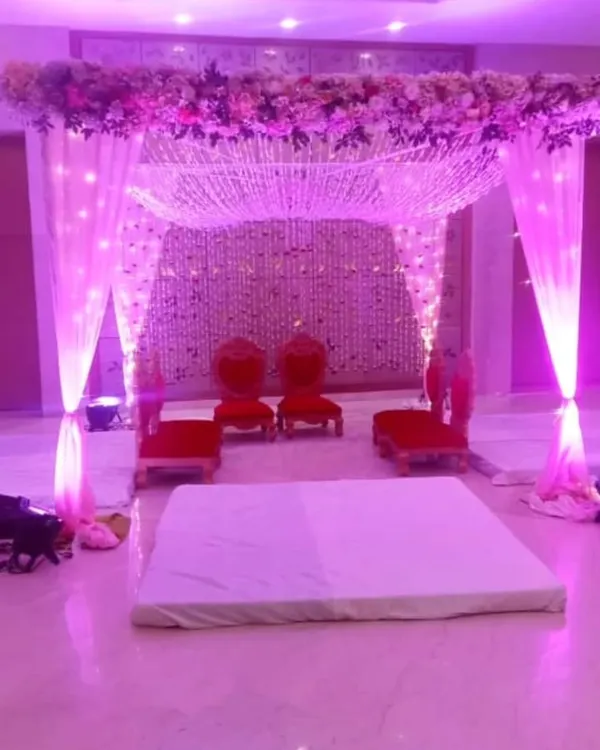 An indoor mandap setup illuminated with beautiful purple lighting. The ceiling is decorated with a web of floral strings, and the seating is arranged for the wedding rituals.