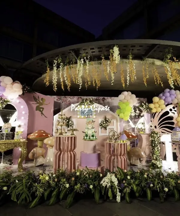 A full view of a fairy and butterfly themed birthday party at dusk. The setup includes a central stage designed like a cottage, flanked by balloon arches, illuminated butterfly wings, and lush floral arrangements at the base.