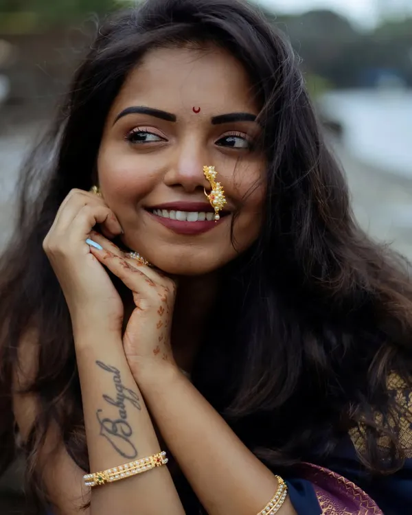 A close-up portrait capturing a joyful, natural smile. The focus is on her expressive face, traditional nath (nose ring), and the 'Babygal' tattoo, showing how modern style and tradition can blend beautifully in a single frame.