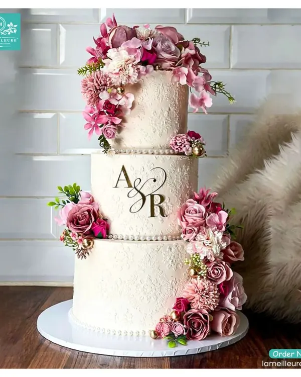 This three-tiered wedding cake features a rustic textured buttercream finish and is personalized with the couple's initials, 'A & R'. The design is elevated by a cascading arrangement of fresh flowers in shades of mauve and deep pink.
