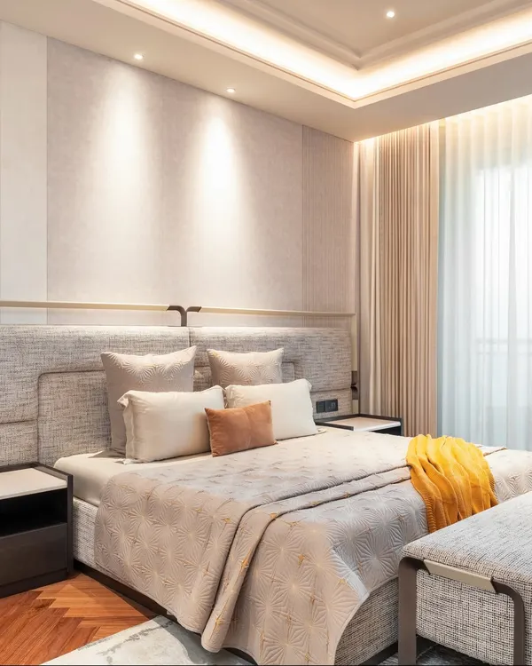 A serene and luxurious master bedroom featuring a custom-designed upholstered headboard that extends across the wall. Integrated side tables and warm, layered lighting create a cohesive and restful environment. The soft textures and neutral palette are accented by a pop of yellow for warmth.