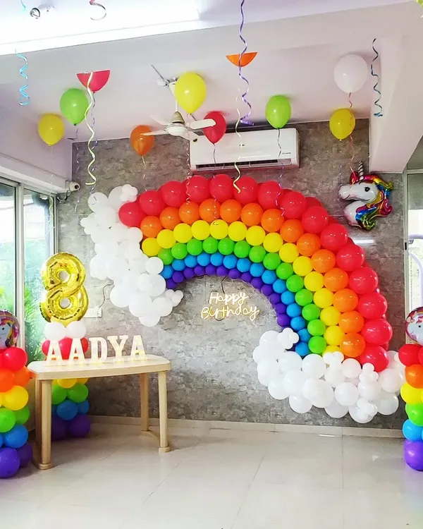 A bright and cheerful rainbow-themed party setup for an eighth birthday. This features a large balloon rainbow arch set against a grey wall, complete with fluffy white balloon clouds, a glowing "Happy Birthday" neon sign, and personalized name blocks for "AADYA". The scene is completed with two rainbow balloon columns and a foil unicorn balloon, creating a magical atmosphere for any kids party.