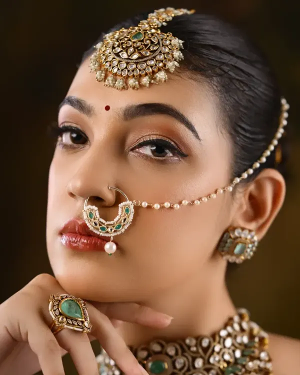A close-up of a North Indian bridal look, focusing on the intricate nath (nose ring) and maang tikka. The eye makeup is sharp and defined, with a glossy lip to balance the look.