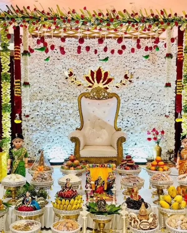 This is the kind of grand, traditional Seemantha setup I love to create. The backdrop is a full wall of fresh white flowers with red accents, and the display includes over a dozen seer plates filled with fruits, sweets, and auspicious items, all centered around the ceremonial chair.
