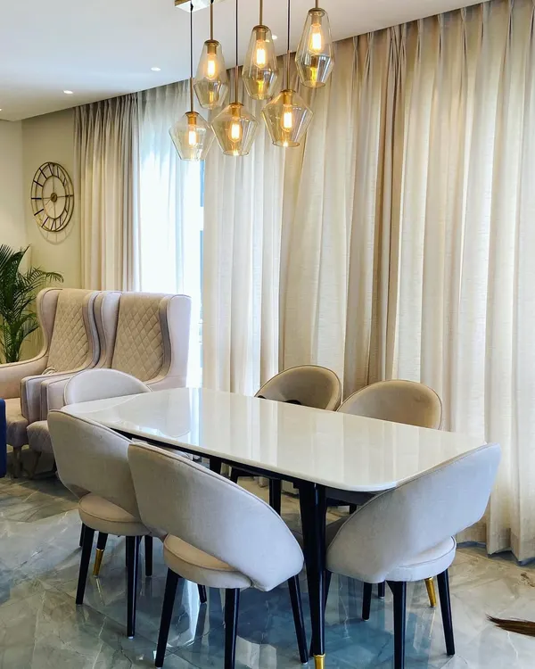 The centerpiece of this dining room in Noida is the stunning cluster of glass pendant lights, which casts a warm glow over the marble-top table. The fully upholstered, curved-back chairs provide exceptional comfort, while the sheer curtains diffuse natural light to create a soft and elegant atmosphere.