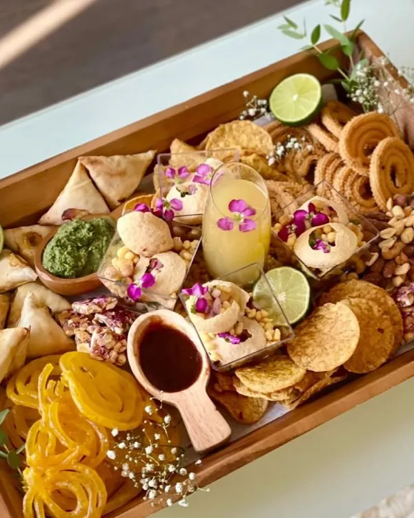 This is my signature desi grazing platter, a feast for the eyes and the palate. I've filled this wooden tray with a mix of classic Indian snacks like samosas, chakli, and jalebi, alongside modern favorites like pani puri shots, all garnished with fresh lime and edible flowers.