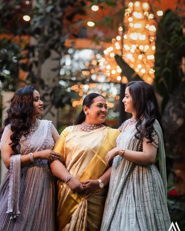 A heartwarming moment between a mother and her two daughters, all dressed in custom Needle Eye creations. The daughters' pastel lehengas beautifully complement their mother's elegant golden saree.