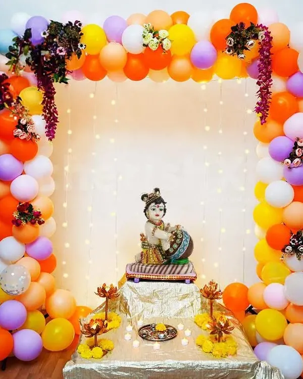 A beautiful setup for a Krishna Janmashtami celebration. A colorful balloon arch frames a small idol of Ladoo Gopal, with fairy lights adding a divine glow.