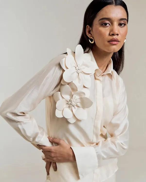 Minimalist structure, maximalist detail. This ivory shirt is elevated with three-dimensional floral appliqués on the shoulder, a true statement of wearable art.