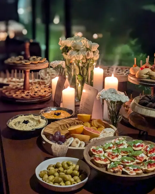 A romantic, candle-lit grazing table perfect for an intimate evening affair. The warm lighting highlights a rich assortment of bruschetta, olives, cheeses, and tiered canapés.