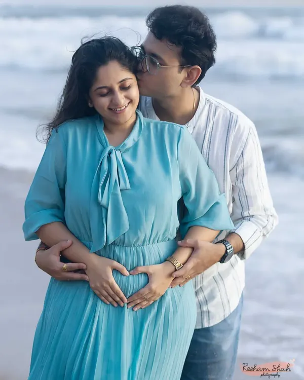 A truly heartwarming moment. The dad-to-be kisses his partner's cheek as they both gently form a heart with their hands on the baby bump, with the ocean waves in the background.