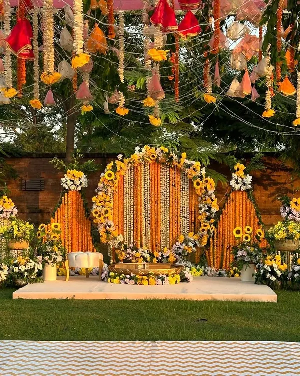 This outdoor Haldi setup is a celebration of sunshine and tradition. A circular stage is framed by pyramids of marigold garlands, creating a vibrant and joyful space for the ceremony.
