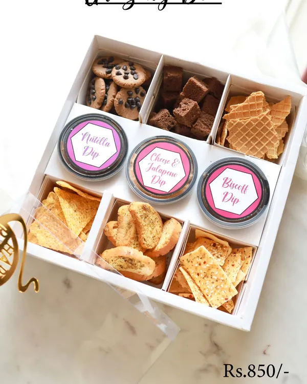 This is our Grazing Box, a perfect Rakhi hamper for sharing. It includes a mix of sweet and savory treats like choco-chip cookies, brownie bites, assorted crackers, and three of our signature dips: Nutella, Cheese & Jalapeno, and Biscoff.