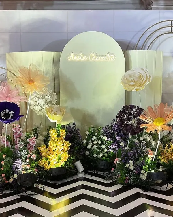 A whimsical garden stage for a cradle ceremony, featuring a neon sign with the baby's name, oversized paper flowers, and a bed of colorful wildflowers. The chevron floor adds a modern, graphic touch.