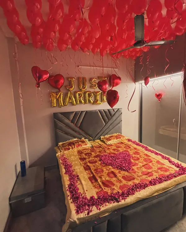 A classic 'Just Married' room decoration. The bed is decorated with rose petals, and the room is filled with red balloons and a foil 'Just Married' banner to welcome the newlyweds.