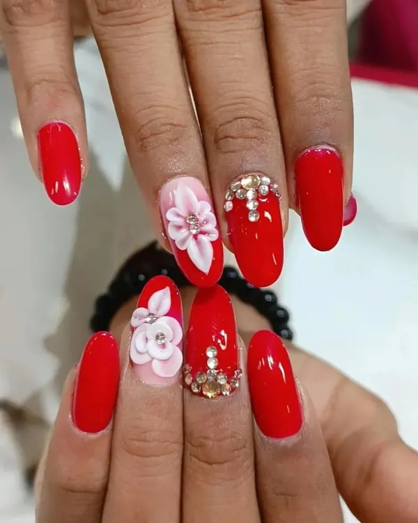 A bold and glamorous set featuring bright red nails, 3D sculpted flowers, and a full accent nail covered in sparkling rhinestones.