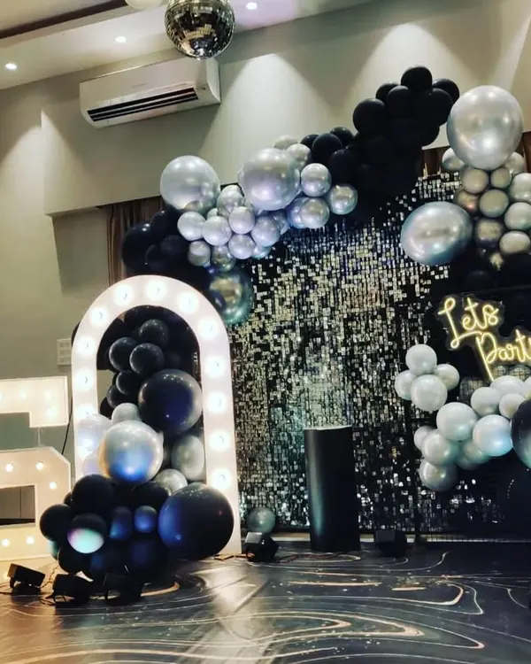 A stylish 25th birthday party setup with a black and silver theme. This decor features a silver shimmer wall, a "Let's Party" neon sign, large '25' marquee numbers, and an elaborate balloon arrangement.