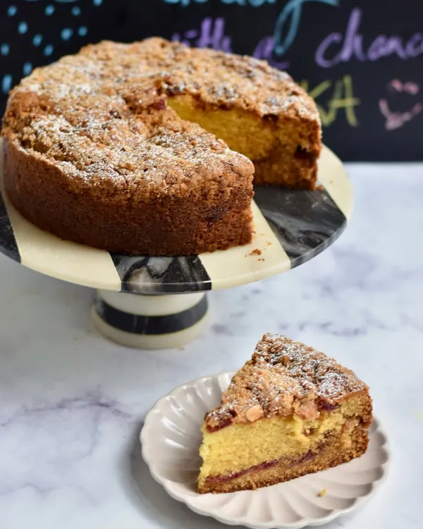 Our Plum & Almond Crumble Tea Cake, with a slice ready to be enjoyed. This comforting cake has a layer of plum jam inside and a crunchy almond crumble on top, perfect for a rainy afternoon with chai.