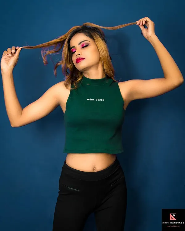 A playful and confident studio portrait. The model, wearing a green crop top, plays with her hair against a solid blue background, creating a vibrant and energetic fashion shot.