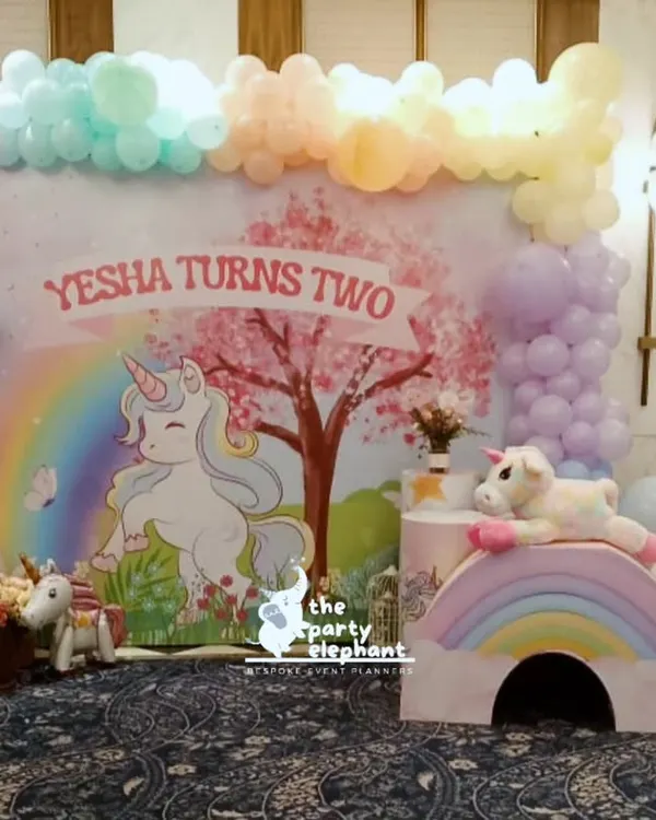 The complete unicorn party setup for Yesha's second birthday. This wide shot shows the main backdrop, pastel balloon garland, rainbow props, and adorable unicorn figures that create a whimsical atmosphere.