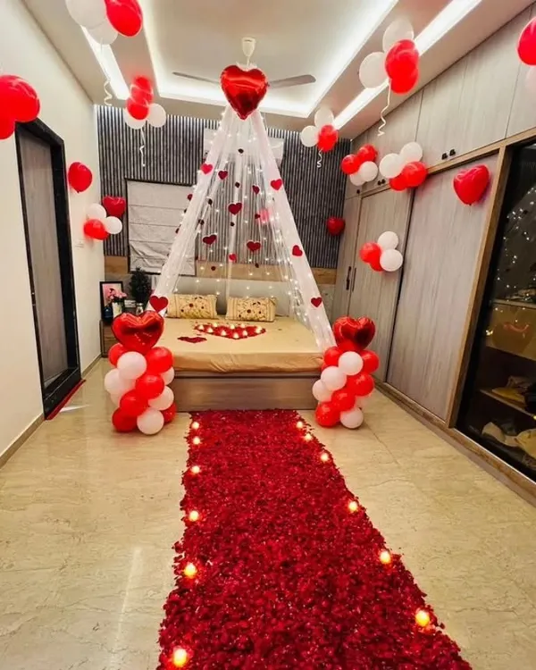 This is my signature romantic room setup. It features a stunning floor path made of real rose petals leading to the bed, which is decorated with a heart shape and a beautiful canopy lit with warm fairy lights. The red and white balloons on the ceiling and floor complete the look, creating an unforgettable surprise for your partner.