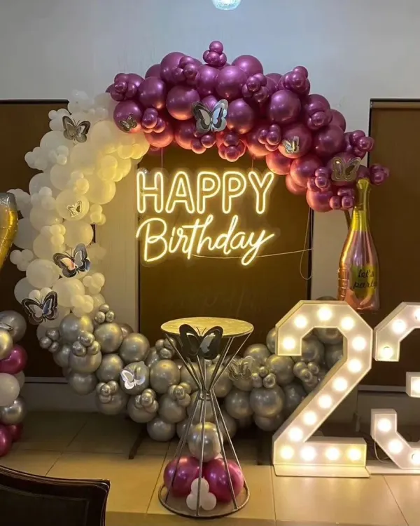 A complete birthday party setup for a 23rd celebration, featuring a bright 'Happy Birthday' neon sign and illuminated marquee numbers. The balloon arch uses a mix of purple, silver, and white, with butterfly props adding a whimsical touch.