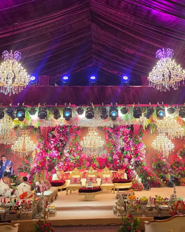 A wide view of a spectacular wedding stage under a tent, illuminated by multiple crystal chandeliers. The entire backdrop is a dense wall of red and pink flowers, creating an opulent and unforgettable setting.