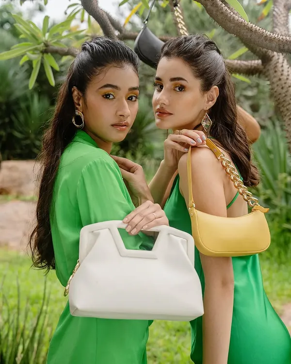 A close-up from the Sarjaa sustainable handbag campaign. This shot highlights the interaction between the models and the products, showcasing two different bag styles in a lush, green environment.