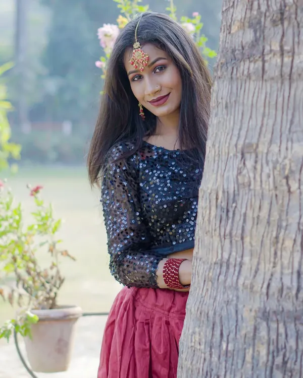 A smiling, candid moment from an outdoor ethnic fashion shoot. The model is peeking from behind a tree, creating a playful yet professional portrait.