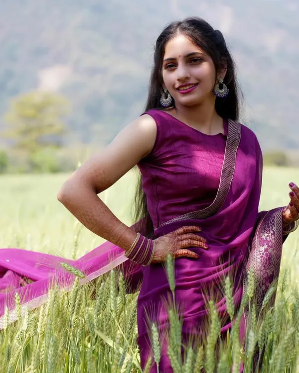 A bright and cheerful outdoor portrait in a field of wheat. The model's confident pose and smile, combined with the flowing purple saree, create a vibrant and lively lifestyle image.