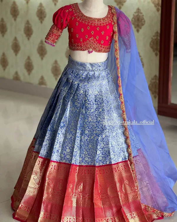 A beautiful kids' lehenga set, featuring a bright pink puff-sleeve blouse with delicate embroidery. The blue brocade skirt has a traditional silk border, and it's completed with a sheer blue dupatta.