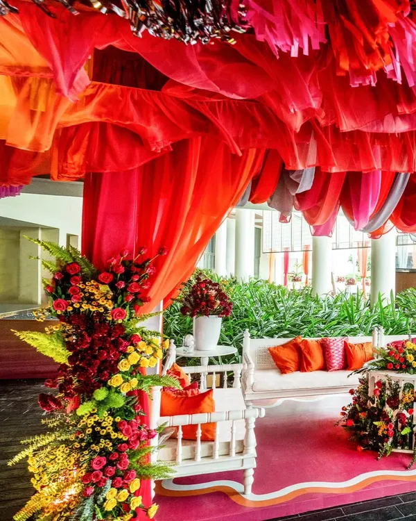 A vibrant Mehendi setting with a canopy of layered red, pink, and orange drapes. The seating area is framed by lush floral pillars, creating a cozy and festive atmosphere.