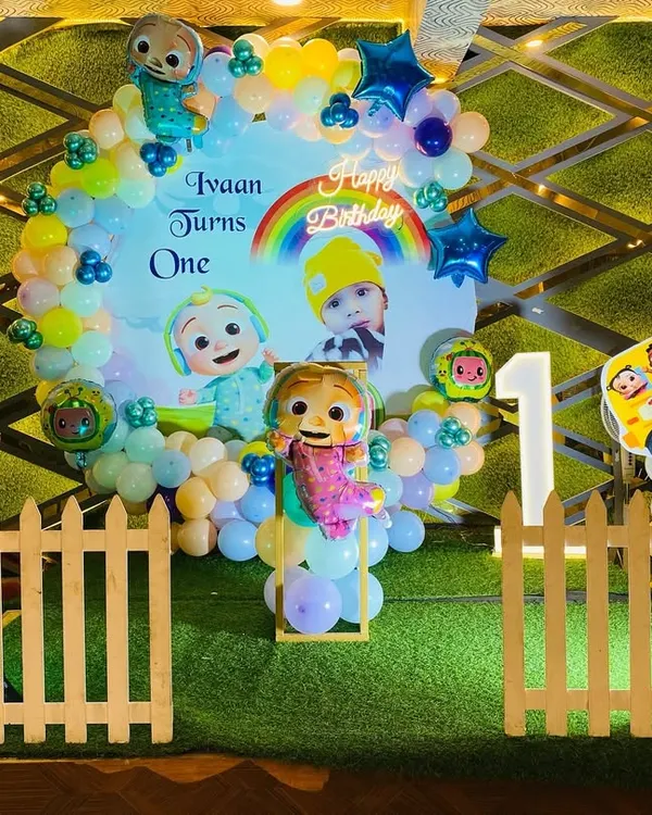 A vibrant Cocomelon theme for a first birthday party. This setup features a custom backdrop with characters, a colorful balloon arrangement, character cutouts, and a picket fence prop to create a fun and immersive experience.