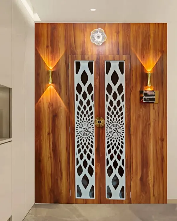The entrance to a home mandir, featuring wooden doors with beautiful white jaali work. The warm wall lights on either side create a welcoming and divine ambiance right at the doorway.
