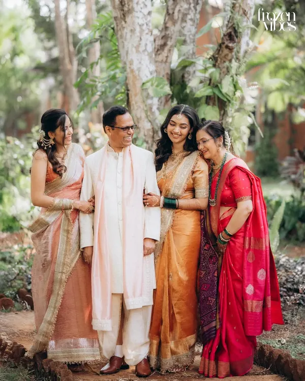 A beautiful family portrait taken in a lush garden setting during a wedding. The bride stands with her father, mother, and sister, all sharing a warm, happy moment together.