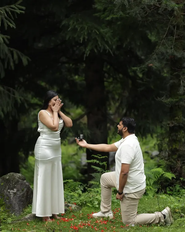 A full view of the surprise proposal, with red petals scattered on the forest floor. This shot captures the entire scene, from his hopeful expression to her genuine shock.