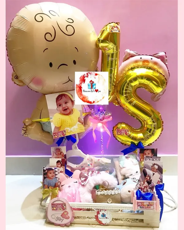 A grand first birthday hamper celebrating a baby girl, complete with "1st" foil balloons, personalized photos, and a crate filled with toys and goodies. This is a wonderful centerpiece gift for a milestone birthday.