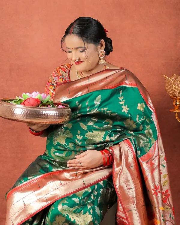 Draped in a magnificent green and red silk saree, this mom-to-be holds a traditional brass thali with flowers, creating a picture of grace and cultural reverence.