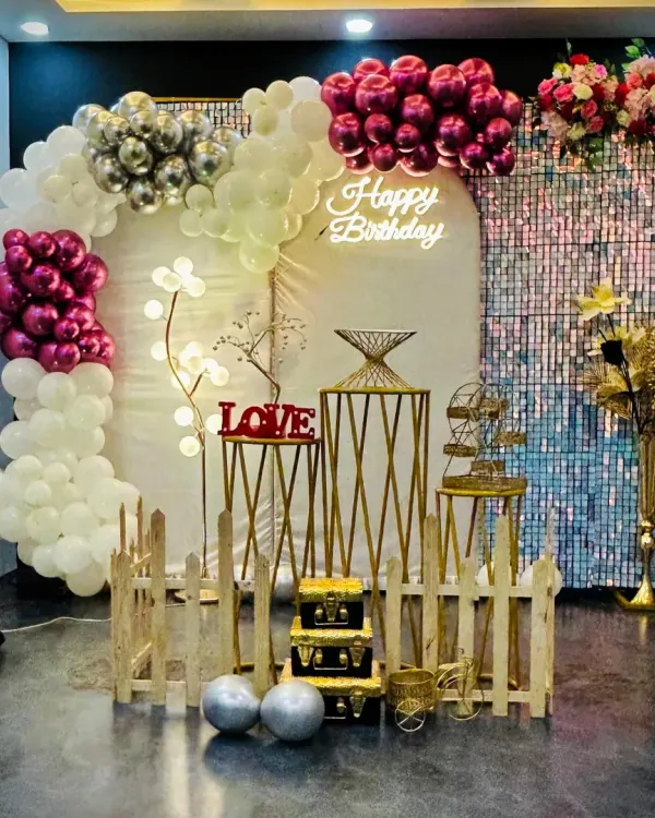 A popular birthday decoration featuring a white, silver, and magenta balloon arch against a shimmering backdrop. The neon "Happy Birthday" sign and decorative pedestals complete the look for a festive party venue.