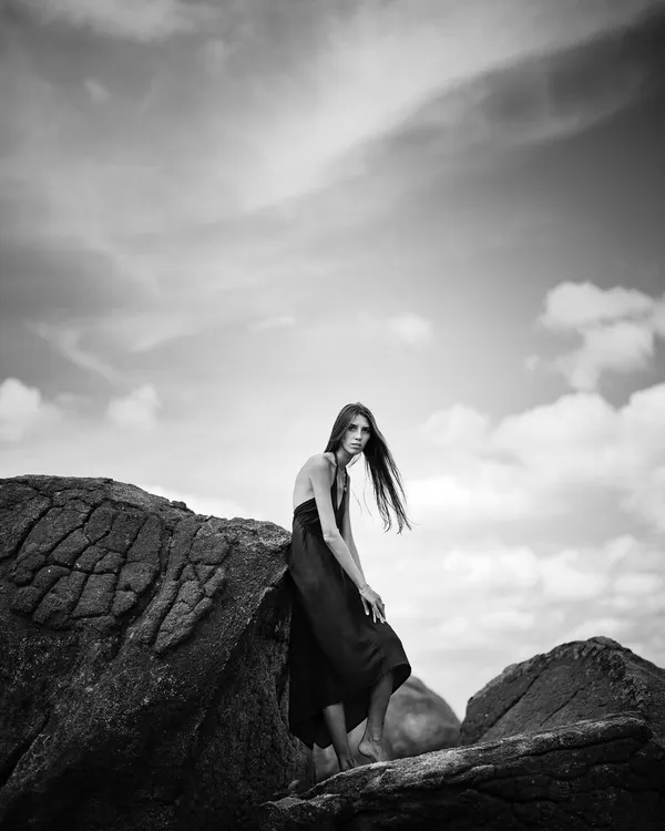 This black and white composition emphasizes the contrast between the dark rocks and the bright, textured sky. The model's pose creates a strong, graphic silhouette.