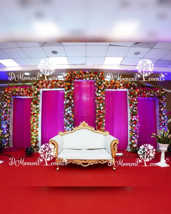 This elegant stage was designed for a 50th golden jubilee anniversary, featuring a classic white and gold loveseat against a backdrop of rich magenta drapes. The archways are heavily decorated with real red, orange, and white flowers, creating a sophisticated and celebratory atmosphere.