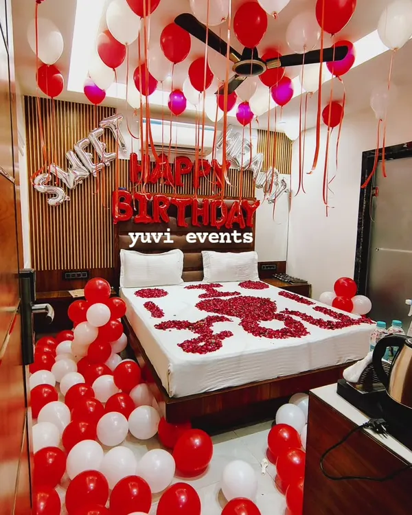 A romantic 'Sweet 16' birthday room surprise. The decoration includes red and white balloons, 'Happy Birthday' foil letters, and a bed decorated with rose petals.