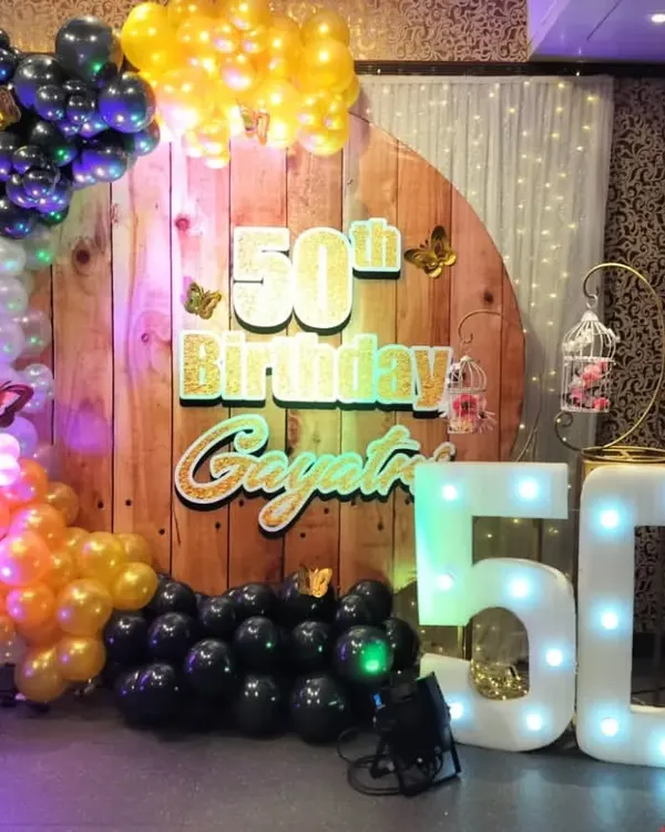 A rustic yet glamorous setup for a 50th birthday. We used a wooden panel backdrop with gold lettering, a multi-colored balloon garland, and a large "50" marquee number to create a unique and celebratory photo corner.